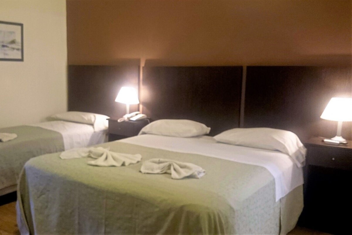 A hotel room with two single beds, each with a pillow and a folded towel on the bed. There is a nightstand with a lamp and a phone on each side of the beds.