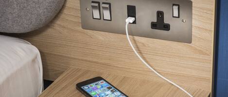 A smartphone charging on a wooden bedside table with a power outlet.