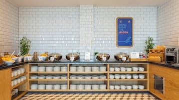 Zona de bufé con diversas opciones de comida, incluyendo cereales, frutas y un cartel que indica la disponibilidad de tocino.