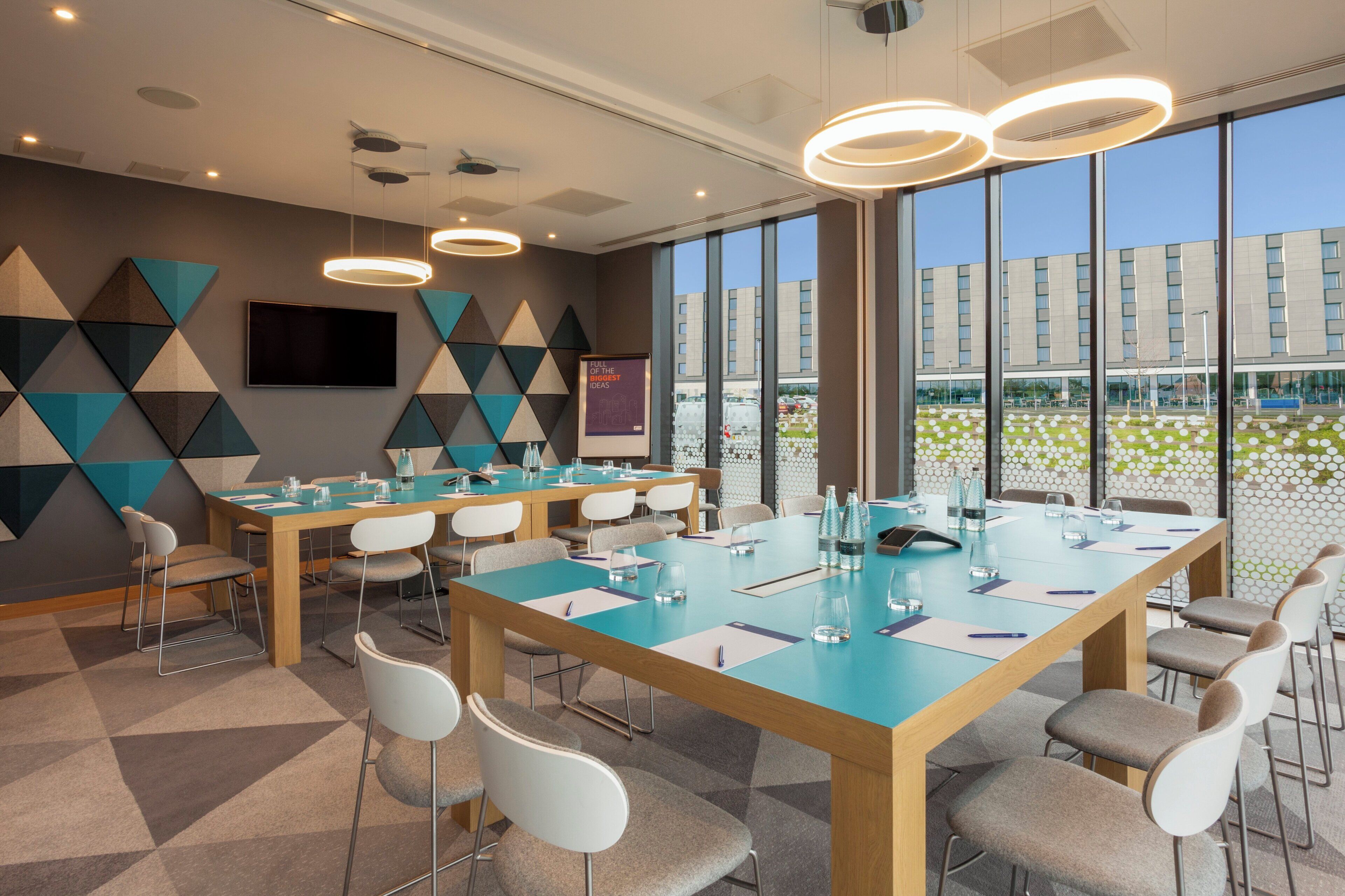 A modern conference room with a large table, chairs, and a large window with a view of a building and greenery.