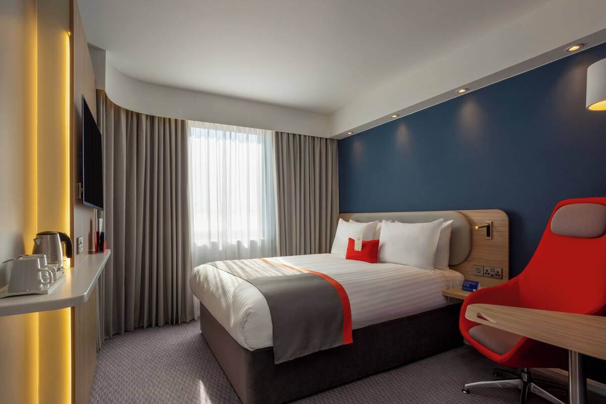 Habitación de hotel con cama, escritorio, silla, televisor y ventana con cortinas.