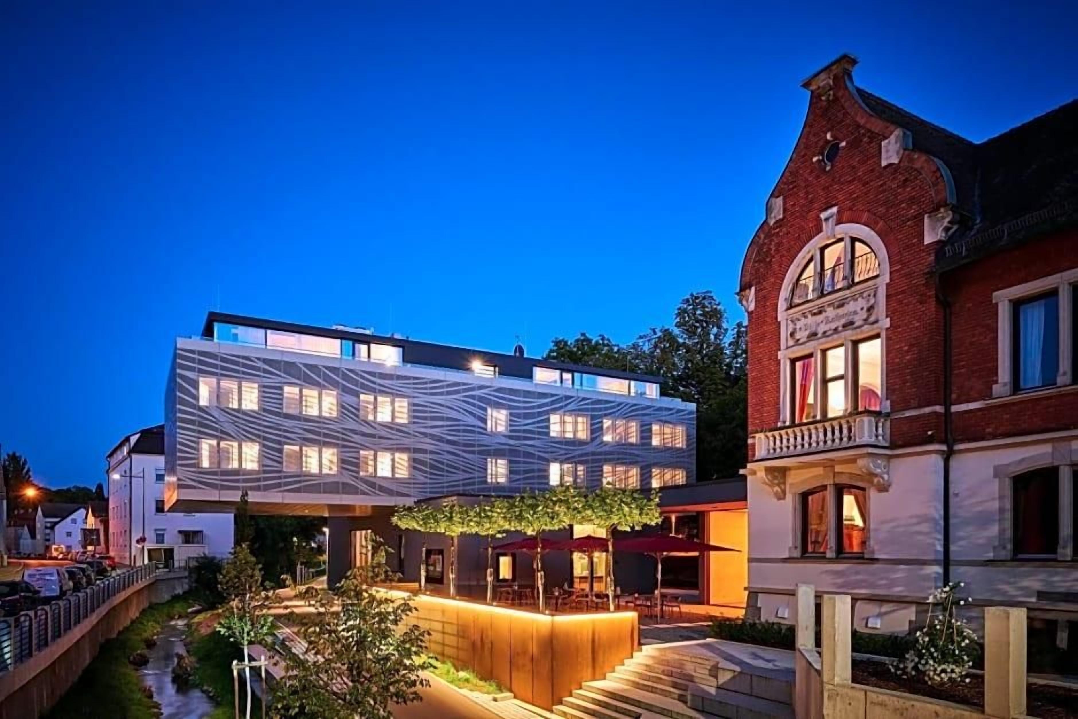 A modern building with a unique geometric facade, a traditional building with a balcony, and a well-lit outdoor area with steps and greenery.