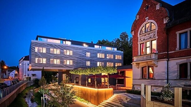 A modern building with a unique geometric facade, a traditional building with a balcony, and a well-lit outdoor area with steps and greenery. - Villa Boutique Hotel (Straubing)