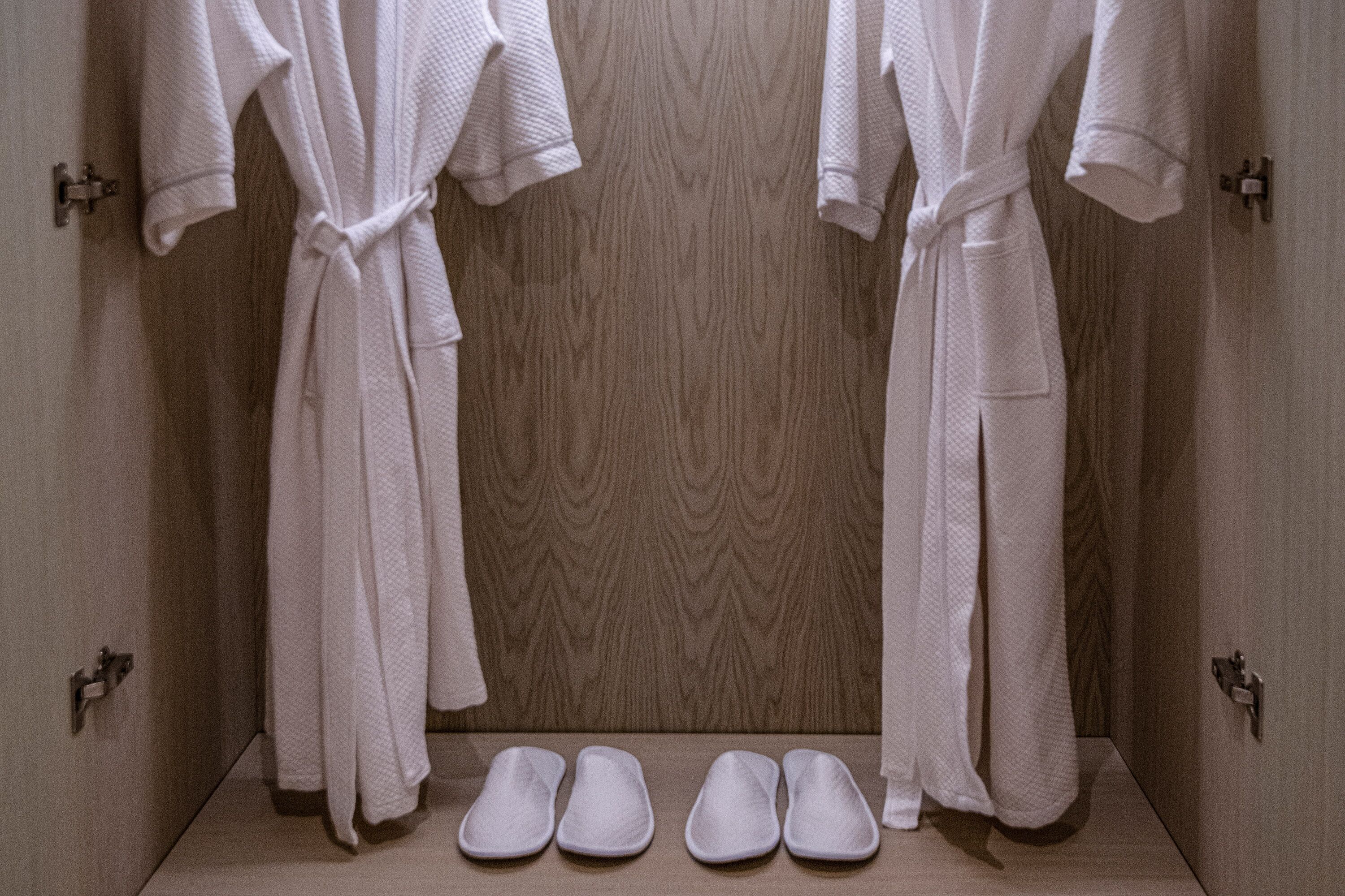 A bathroom with wooden walls, two white robes hanging on hooks, and four pairs of white slippers on the floor.