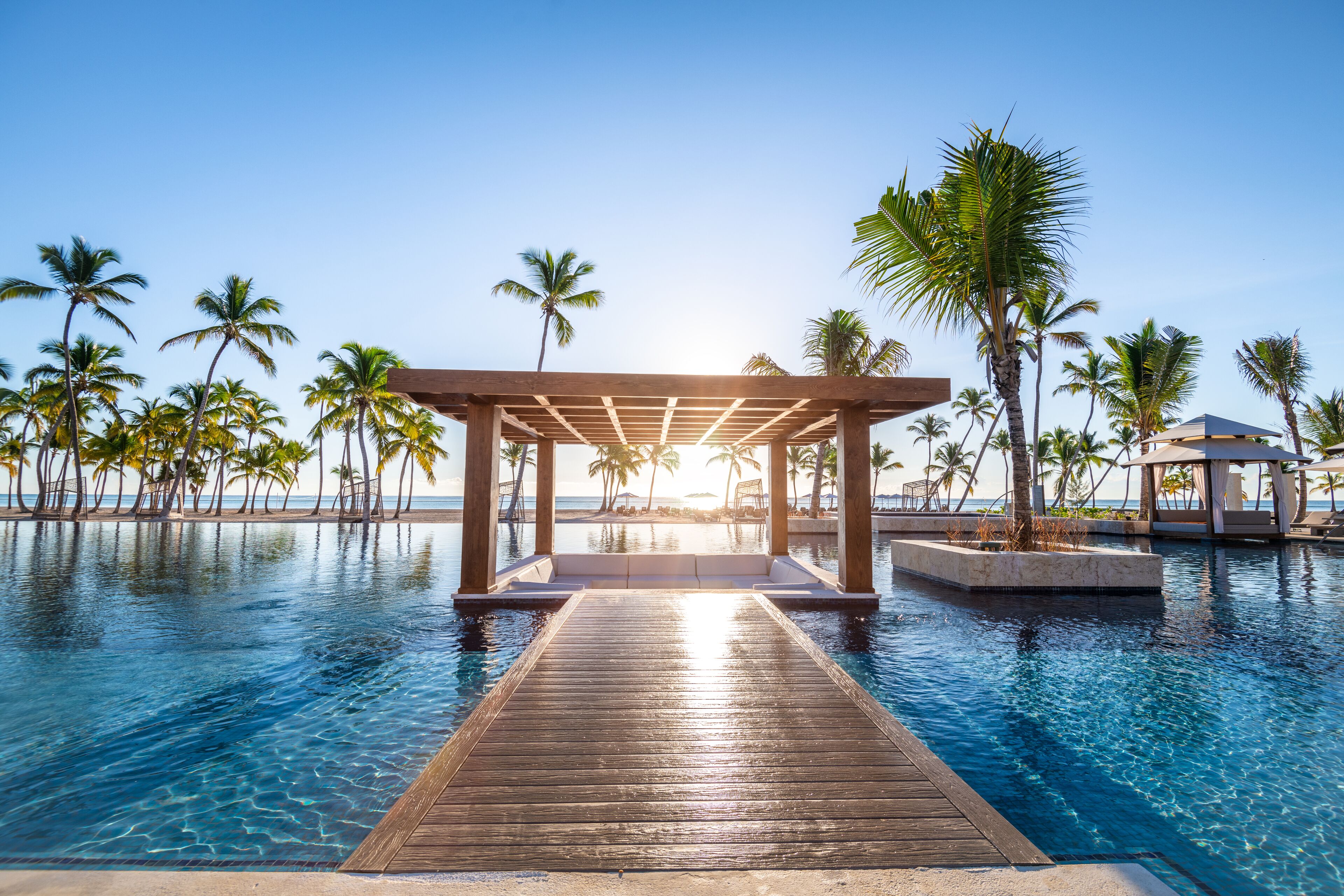 Un pontile in legno che conduce a una piscina, con palme e un cielo sereno.