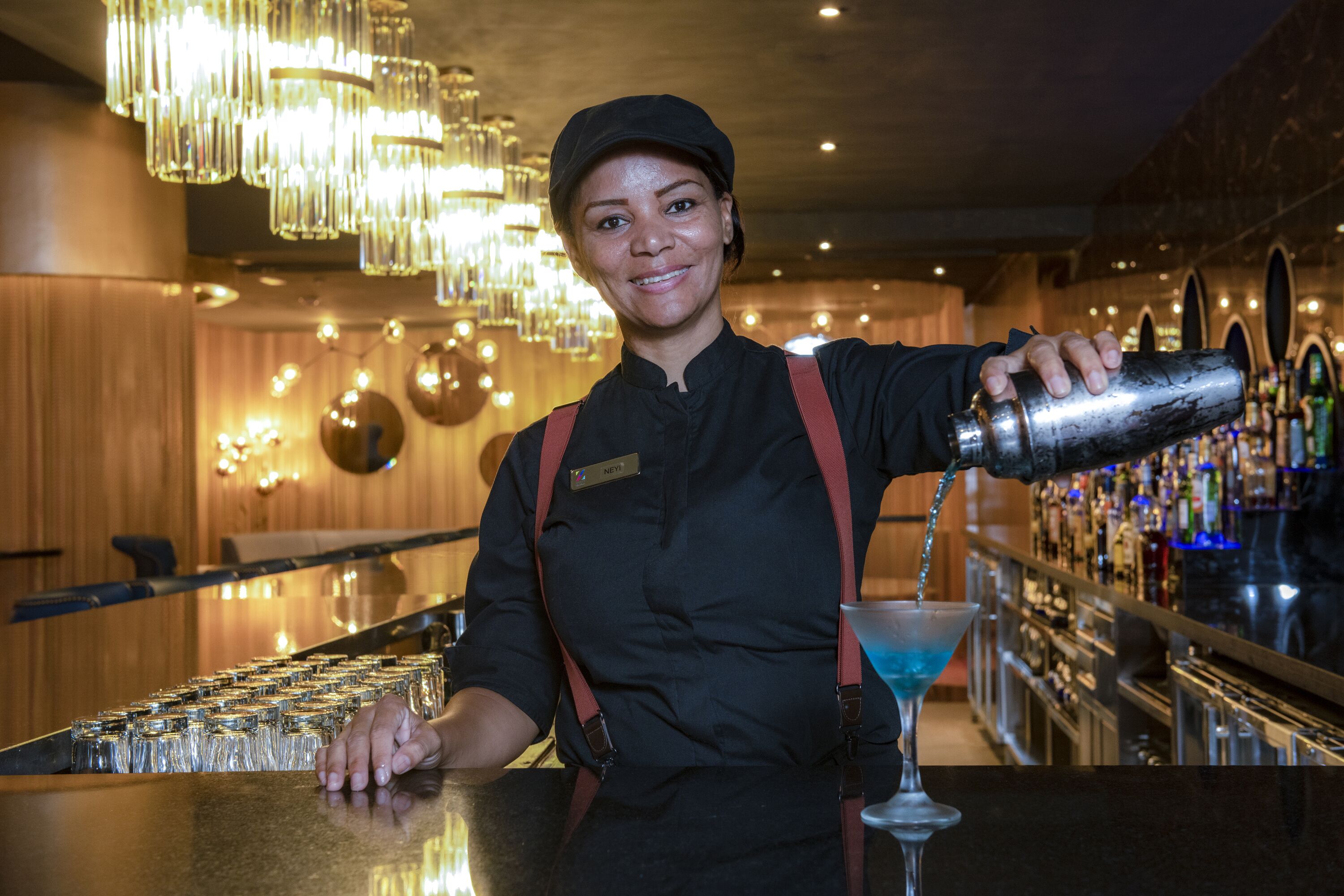 A bartender pouring a drink into a martini glass at a bar with a wooden interior and a chandelier.