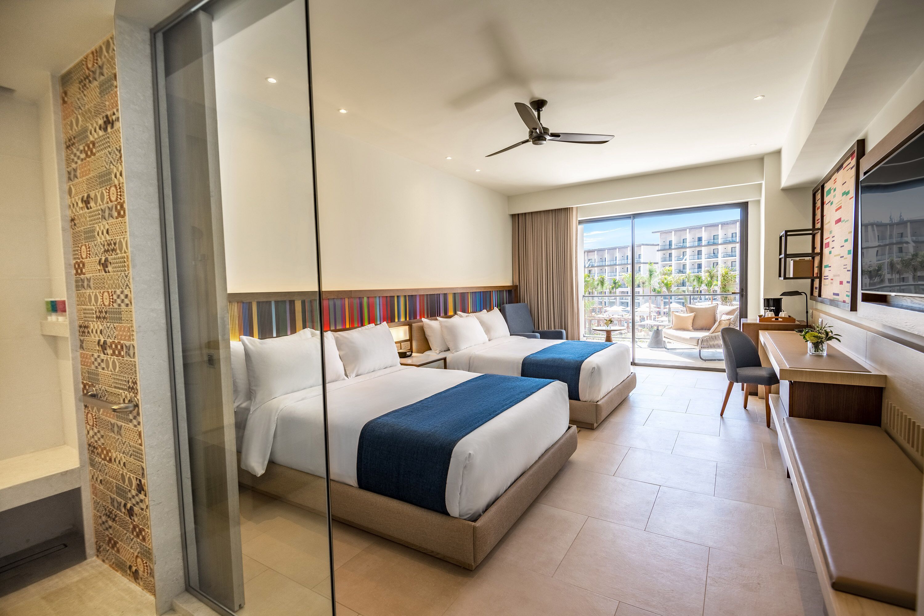 A hotel room with two beds, a desk, a chair, a TV, and a balcony with a city view.