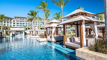 A resort pool area with cabanas, palm trees, and a large building in the background.