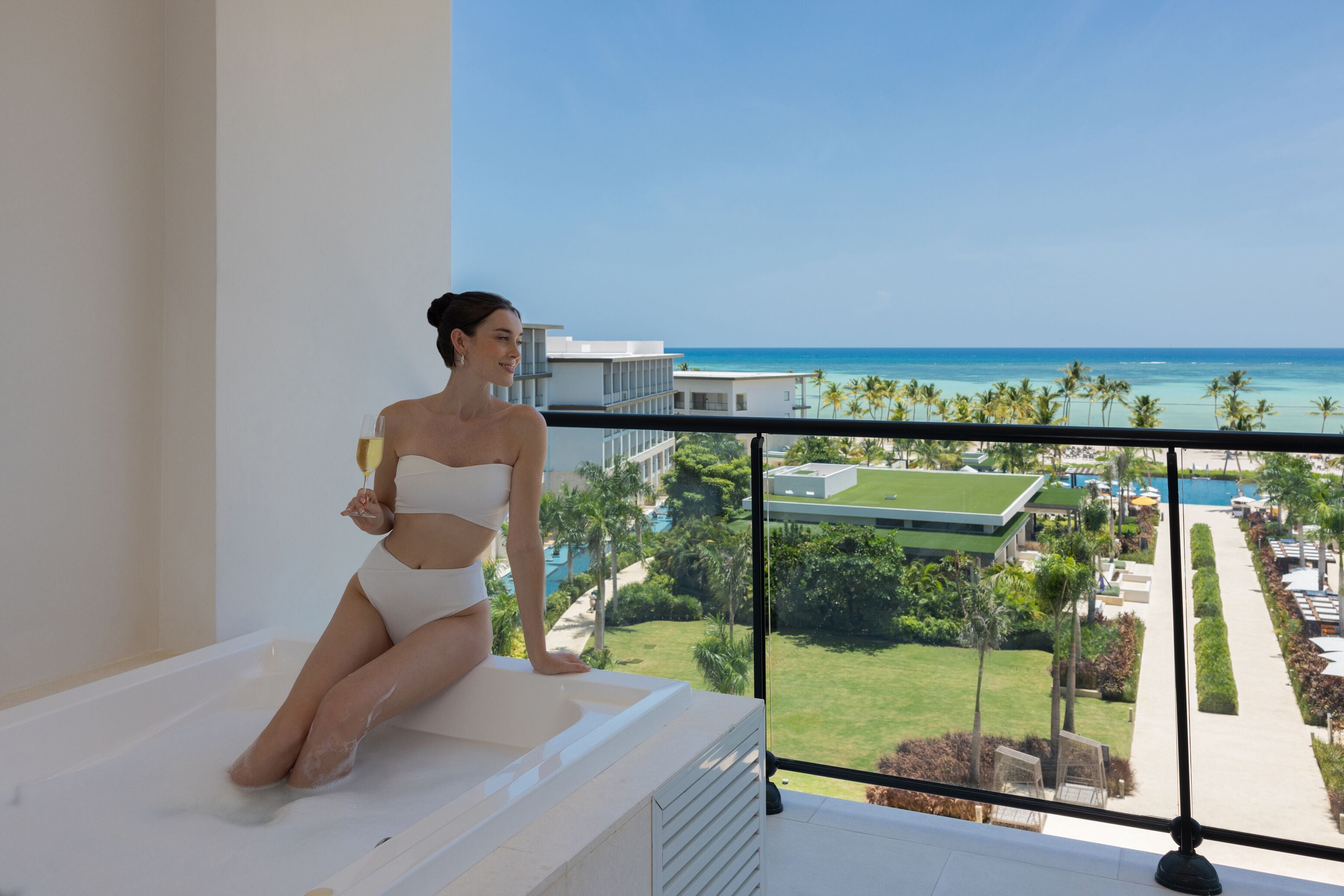 Eine Frau in einer Badewanne auf einem Balkon mit Blick auf einen Pool und Palmen.