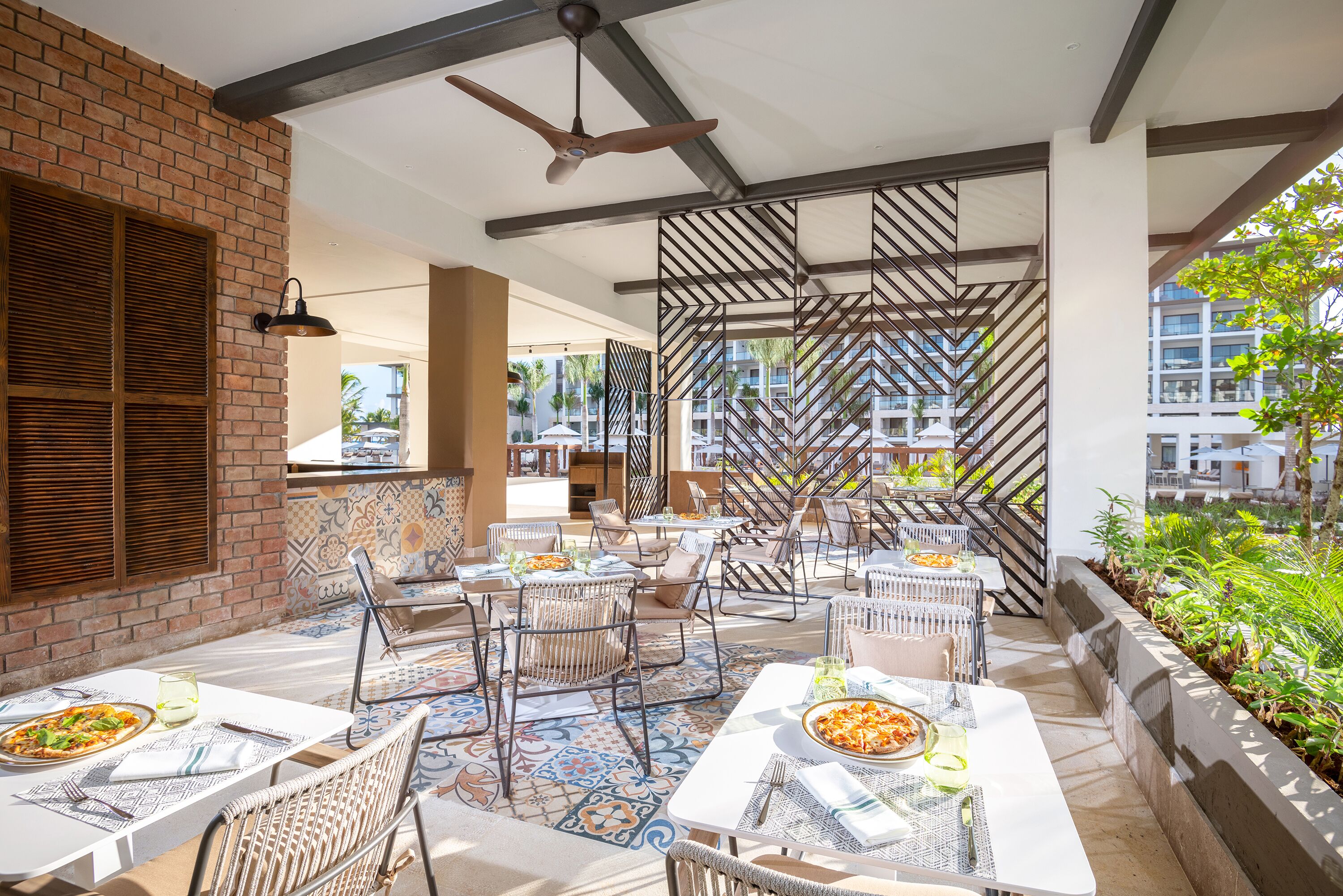Un espace de restauration en plein air doté de plusieurs tables et chaises, d’un ventilateur de plafond et offrant une vue sur les bâtiments environnants.