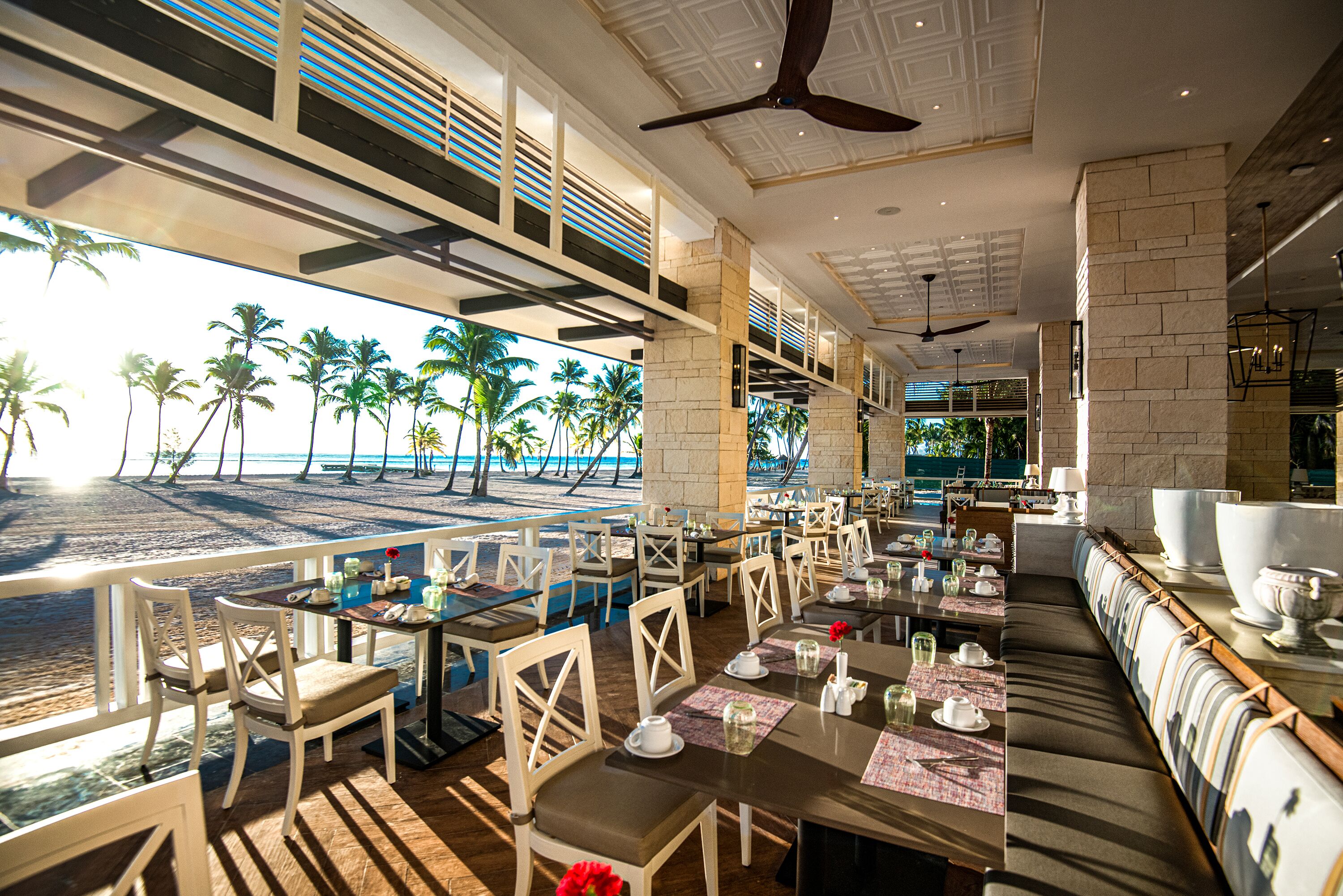 A beachfront restaurant with outdoor seating overlooking the ocean, featuring modern decor and a view of palm trees.