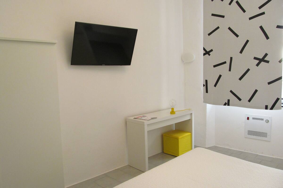 Una habitación de hotel moderna con un televisor de pantalla plana montado en la pared, un escritorio pequeño blanco y un taburete amarillo, y una cama con cabecera blanca.