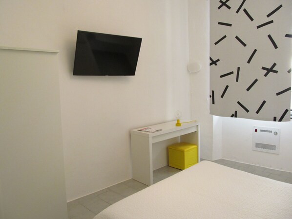 Een moderne hotelkamer met een flatscreen televisie aan de wand, een klein wit bureau met een gele kruk, en een bed met een wit hoofdeinde.