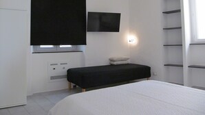 Een moderne slaapkamer met een bed, een zwarte bankje, een aan de muur bevestigde televisie en een raam met een plank.