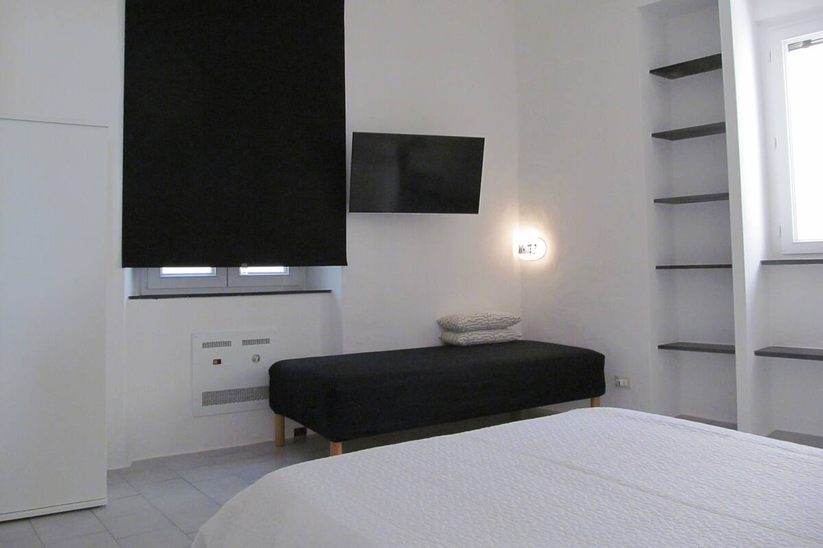 Un dormitorio moderno con una cama, un banco negro, un televisor montado en la pared y un ventanal con estante.