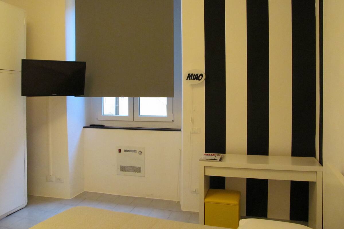 Una habitación con una cama, un televisor montado en la pared, una ventana con persianas, una mesita con una revista y un taburete amarillo.