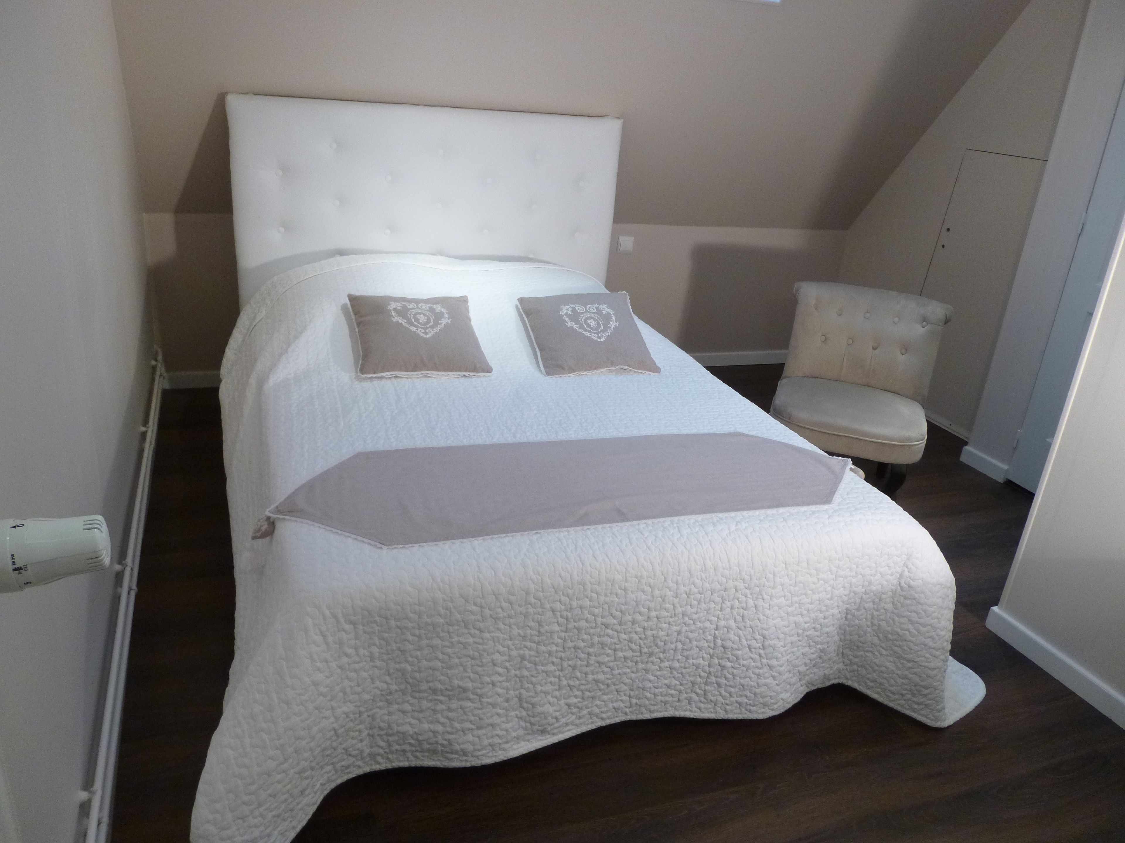 Un lit bien fait, recouvert d’une couette blanche et agrémenté de deux coussins décoratifs, une petite chaise rembourrée et un radiateur fixé au mur.