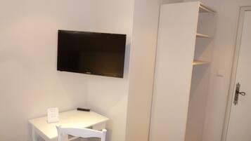 Een kleine kamer met een wit bureau, een stoel, een flatscreen televisie aan de muur en een witte garderobe met open planken.