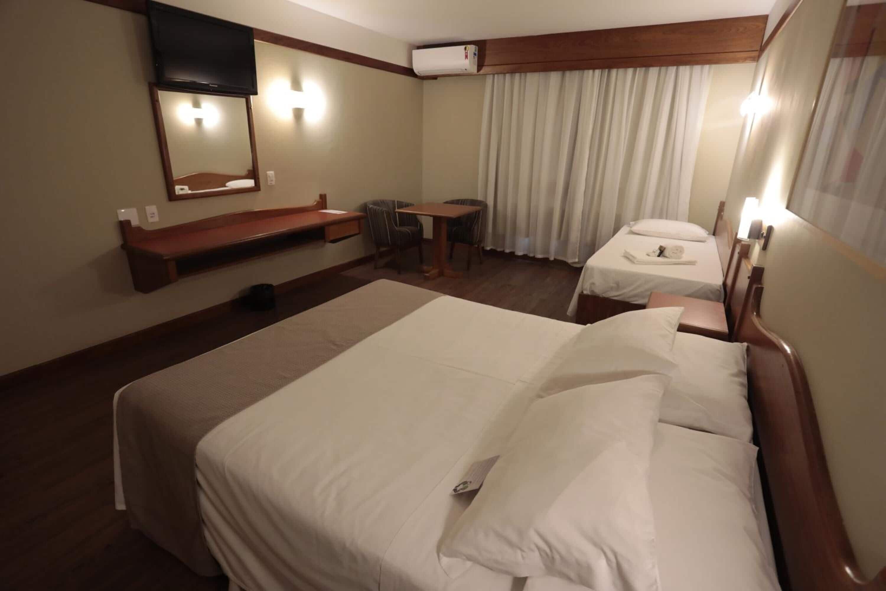 A hotel room with a large bed, a smaller bed, a desk, a chair, a mirror, and a television.