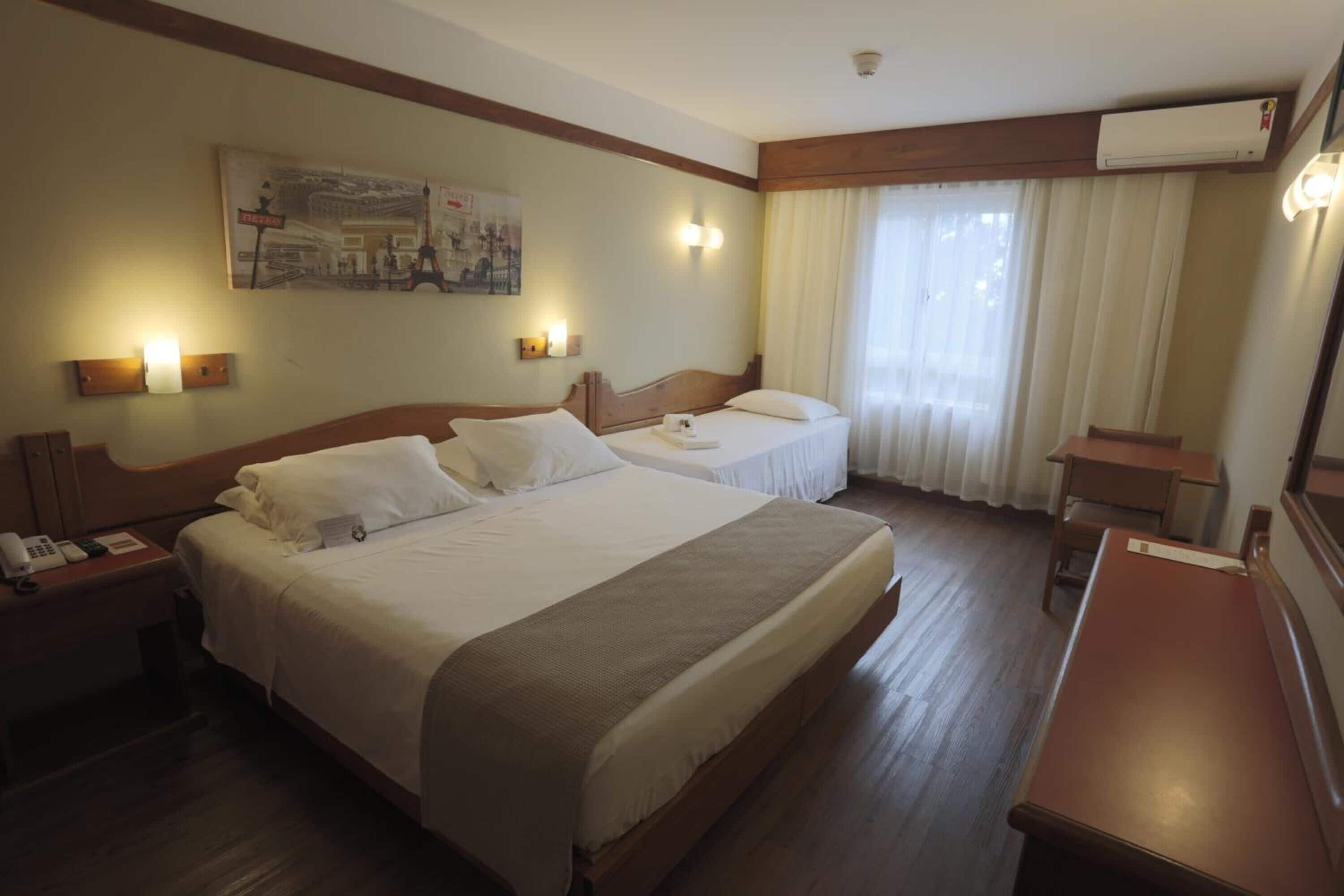 A hotel room with two beds, a desk, and a chair. There are wall-mounted lights and an air conditioning unit.