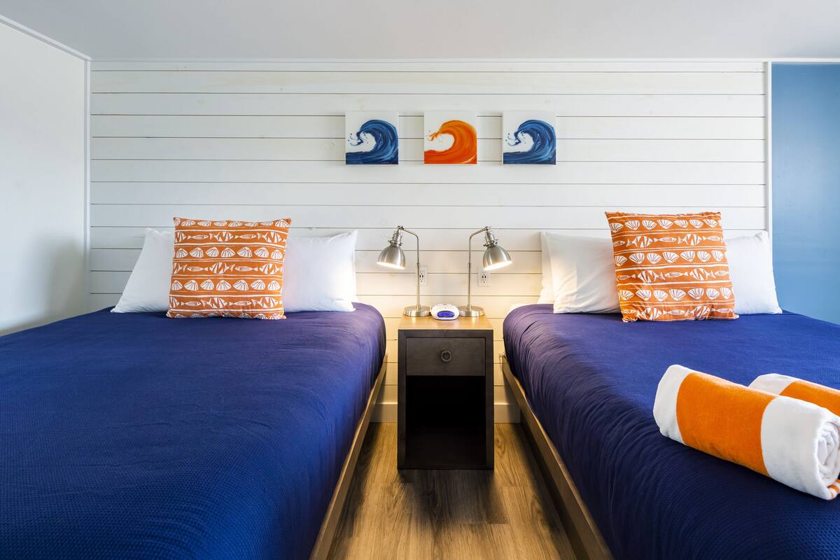A hotel room with two beds, each with matching blue bedding and orange pillows with a shell pattern. There are wall-mounted lamps on either side of the beds, and a small table between them.