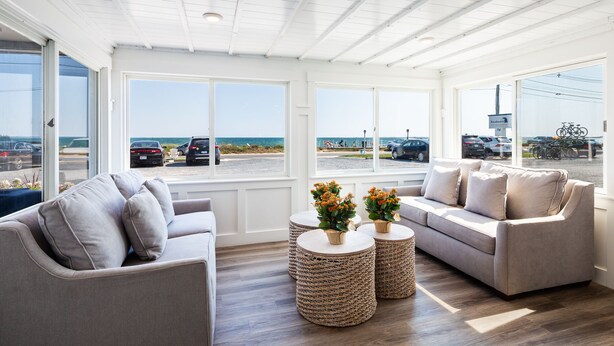 A spacious living room with a large window offering a view of the beach and parked cars. There are two sofas and two round woven coffee tables with flower arrangements. - Breakwater Hotel (Provincetown)