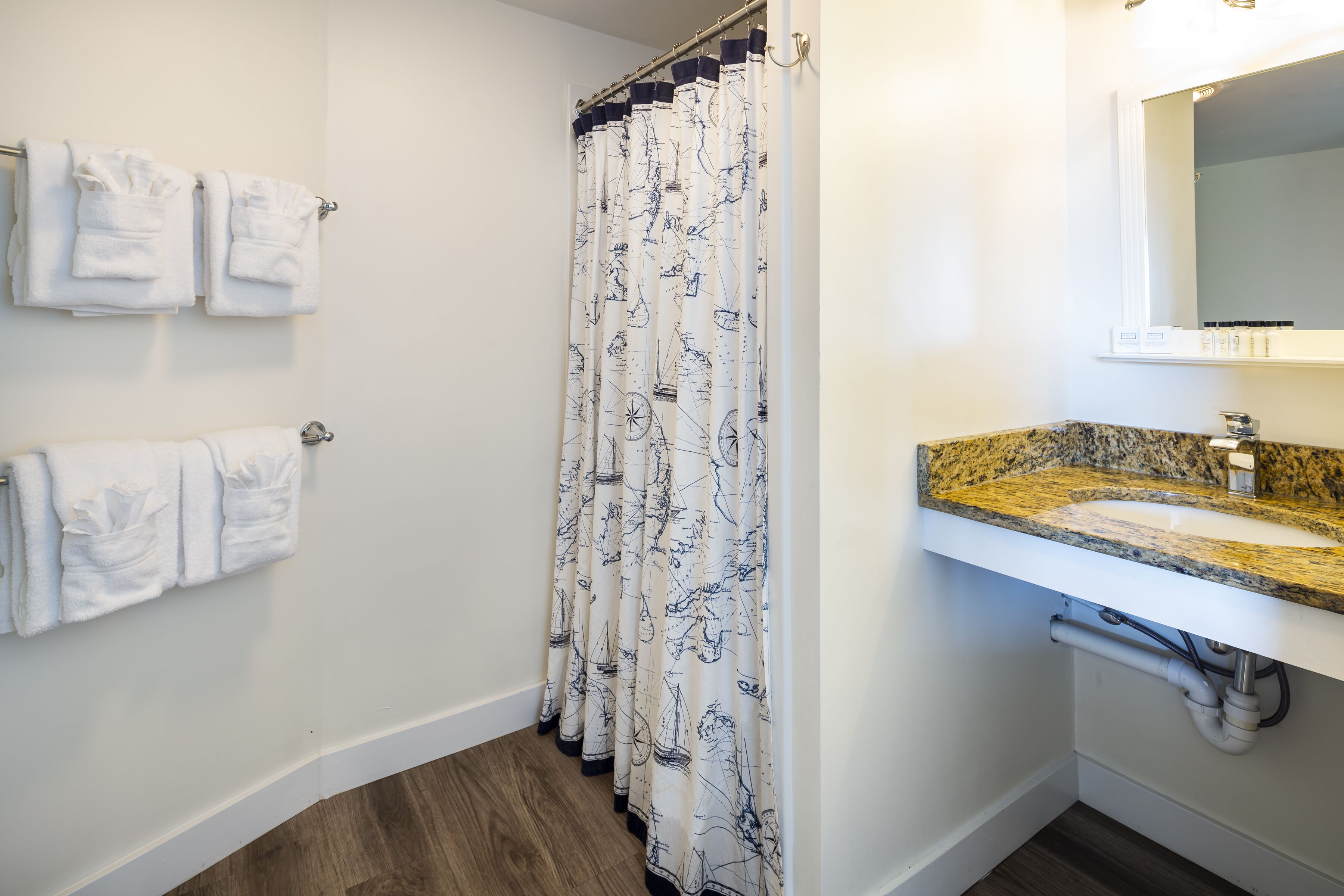 A bathroom with a shower curtain featuring nautical designs, a sink with a granite countertop, and multiple white towels hung on the wall.