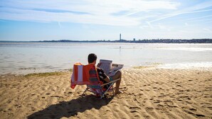 A person sitting on a beach chair with a newspaper, facing a calm sea with a distant cityscape. - Breakwater Hotel (Provincetown)