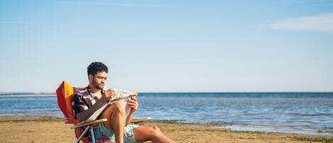 Eine Person sitzt am Strand auf einem Liegestuhl und liest ein Buch.