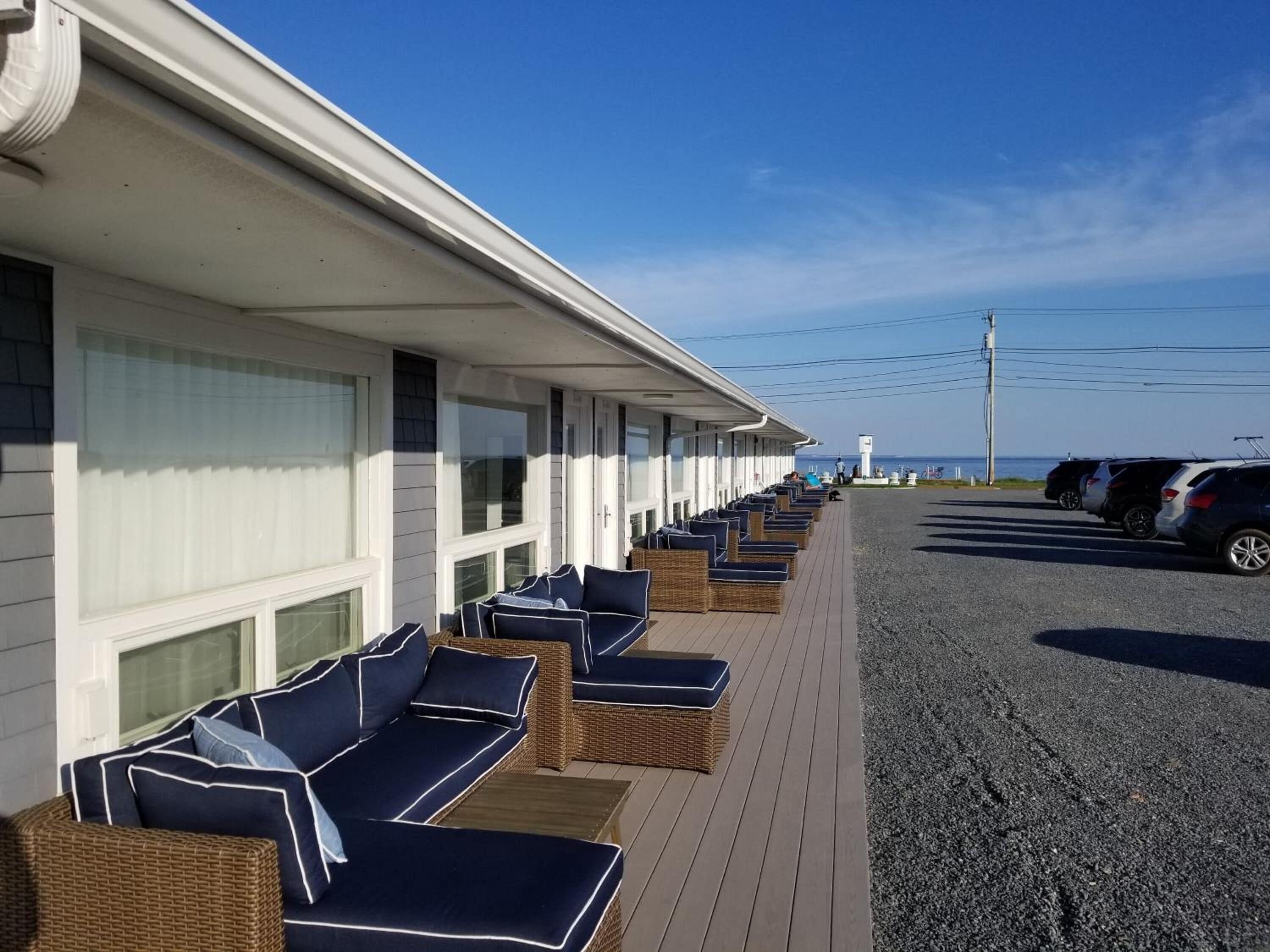 A row of wicker lounge chairs with cushions, aligned along a covered walkway adjacent to a building with large windows.