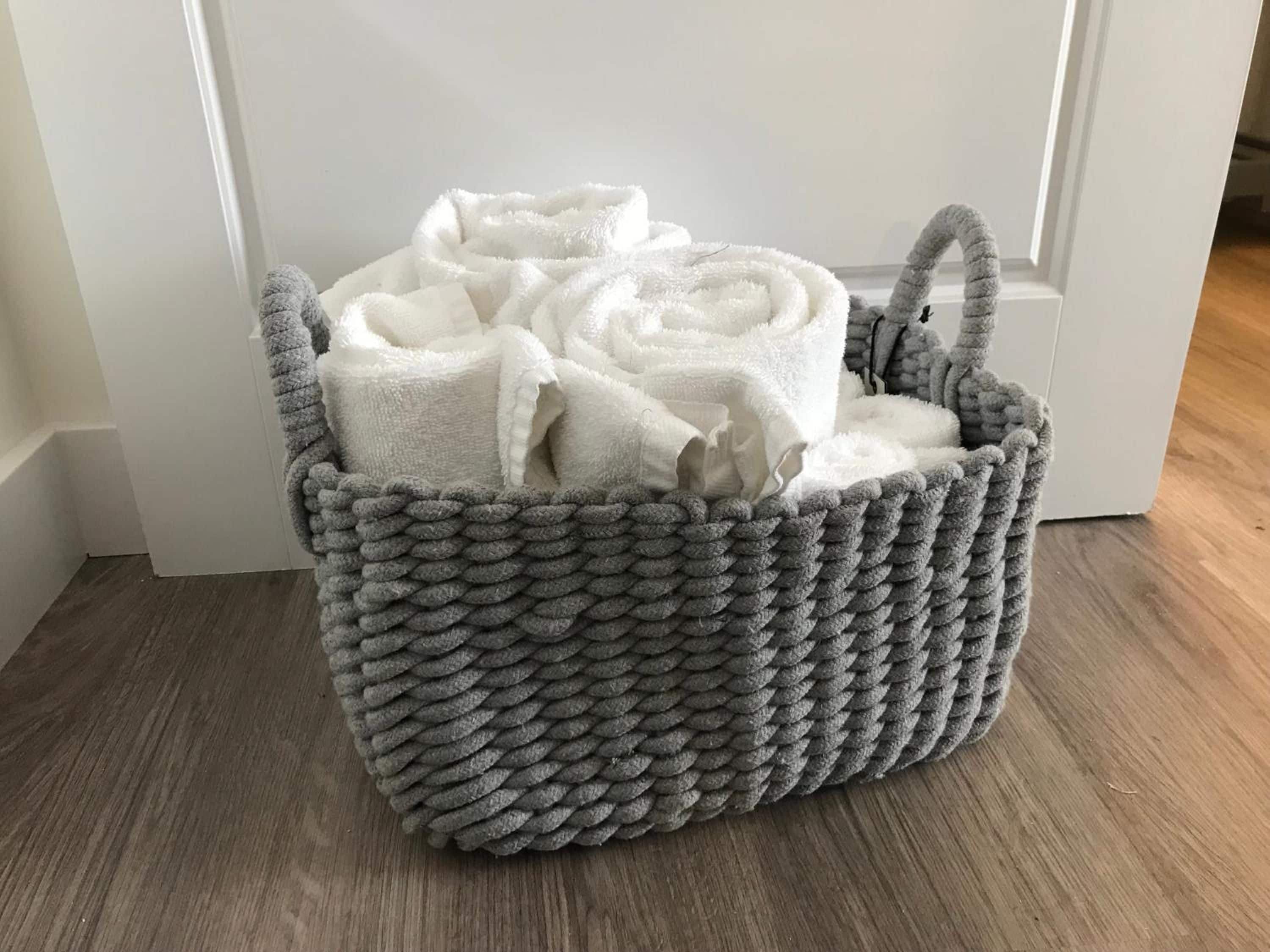 Un cesto grigio intrecciato pieno di asciugamani bianchi su un pavimento di legno, di fronte a una porta bianca.