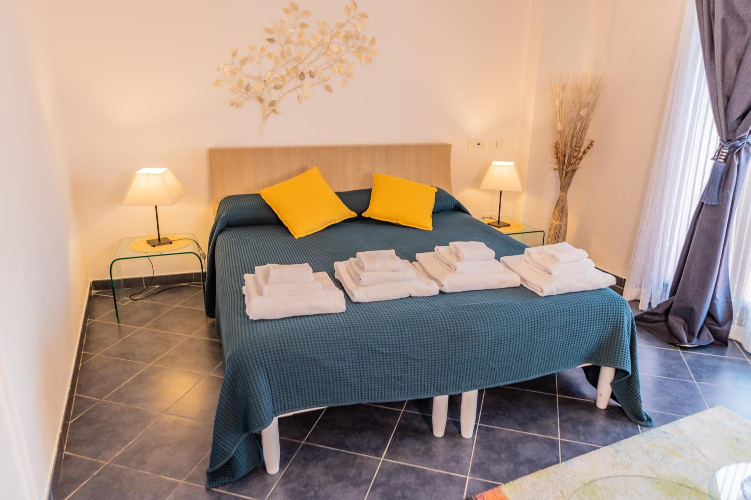 Een net opgemaakt bed met een blauwgroene sprei, twee heldergele kussens en een stapel witte handdoeken op het voeteneinde.