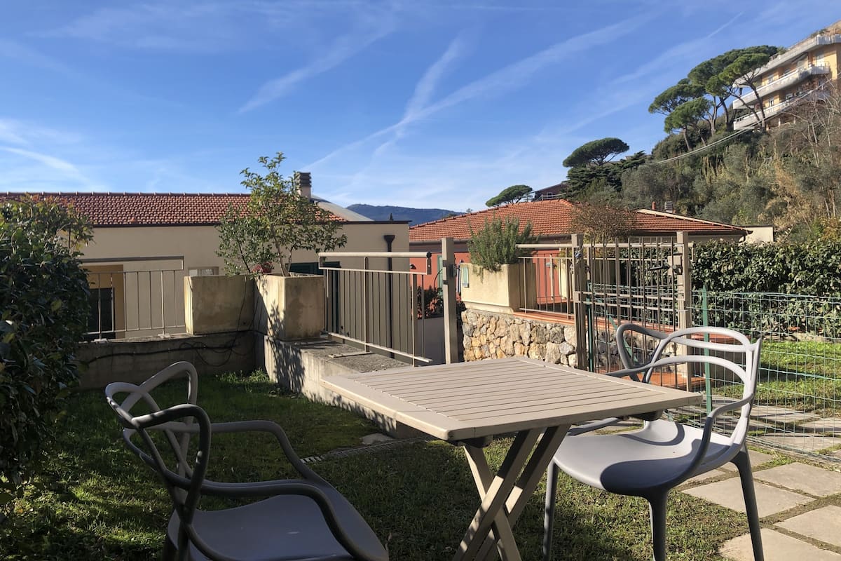 Un patio al aire libre con una mesa y sillas, rodeado de un muro de piedra y vegetación, con un edificio al fondo.