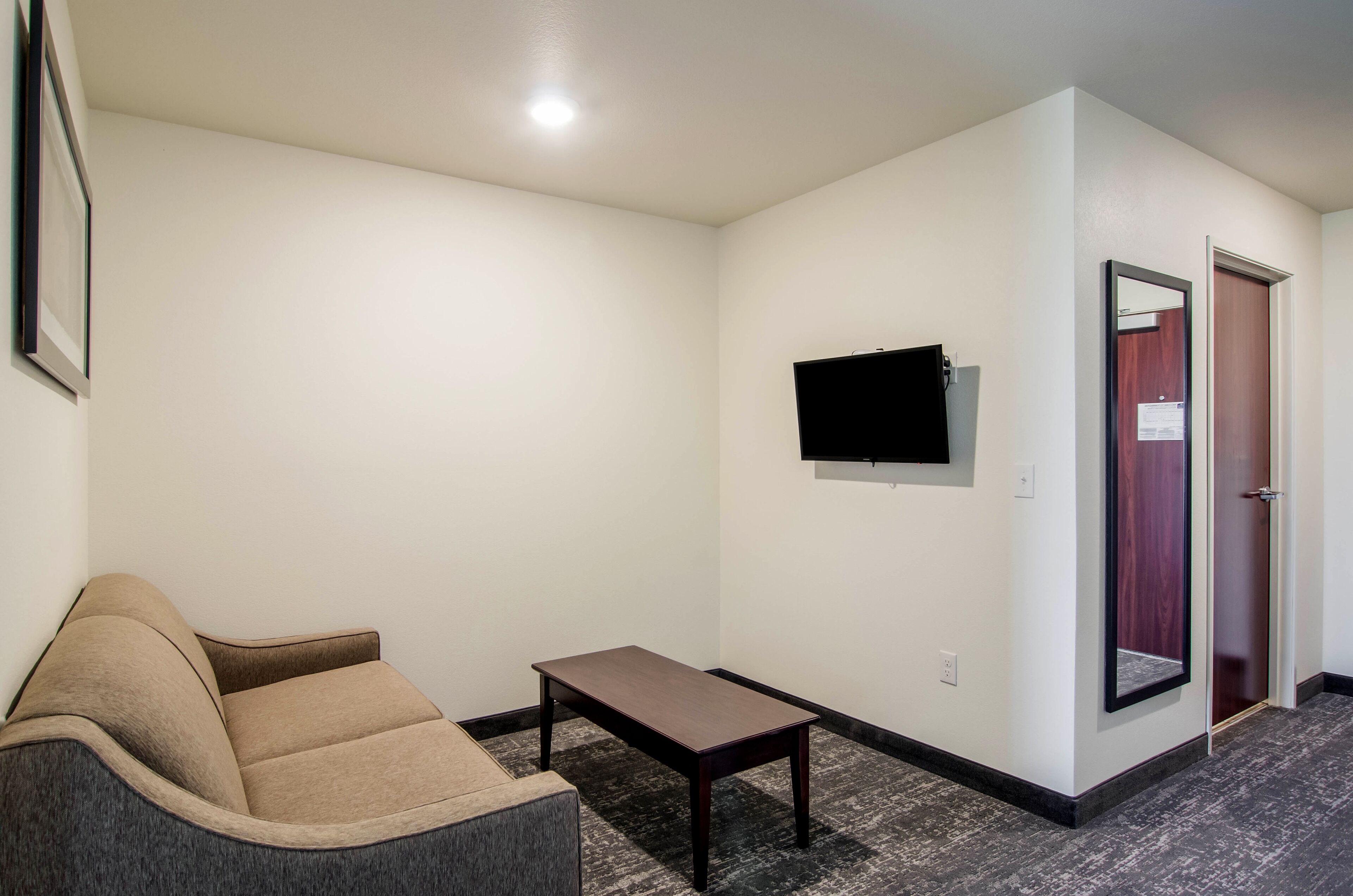 A hotel room with a sofa, a small table, a flat-screen TV mounted on the wall, and a door with a mirror.