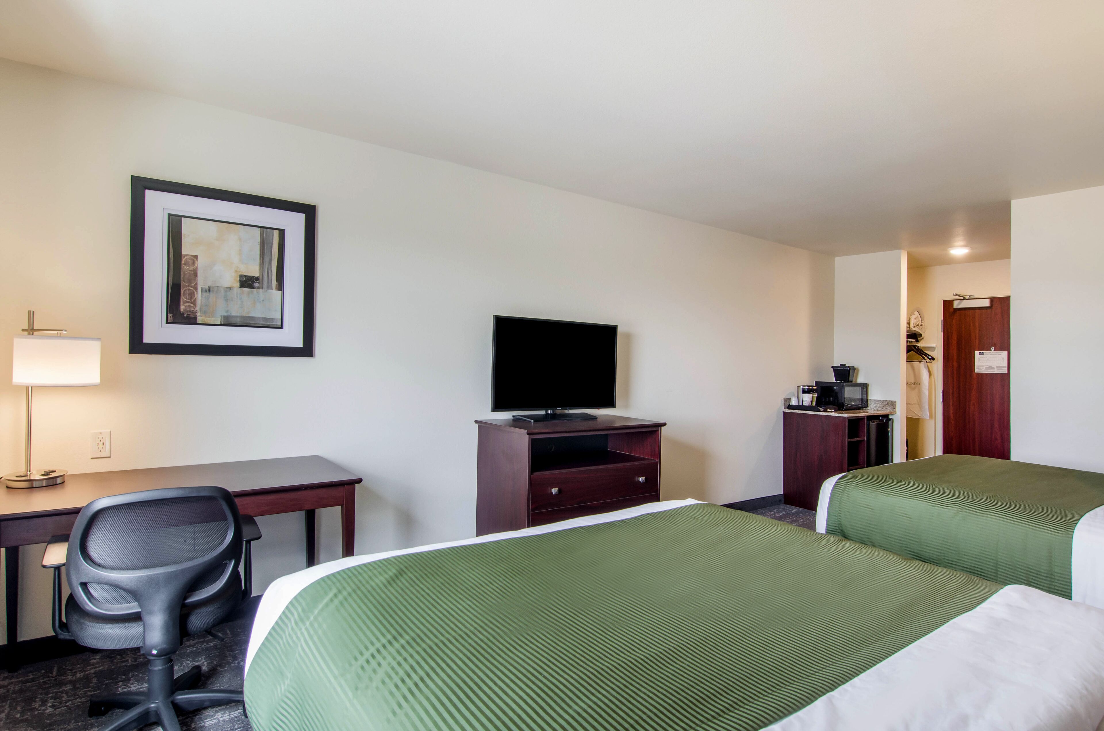 Ein Hotelzimmer mit zwei Betten, einem Schreibtisch, einem Sessel, einem Fernseher und einem gerahmten Bild an der Wand.