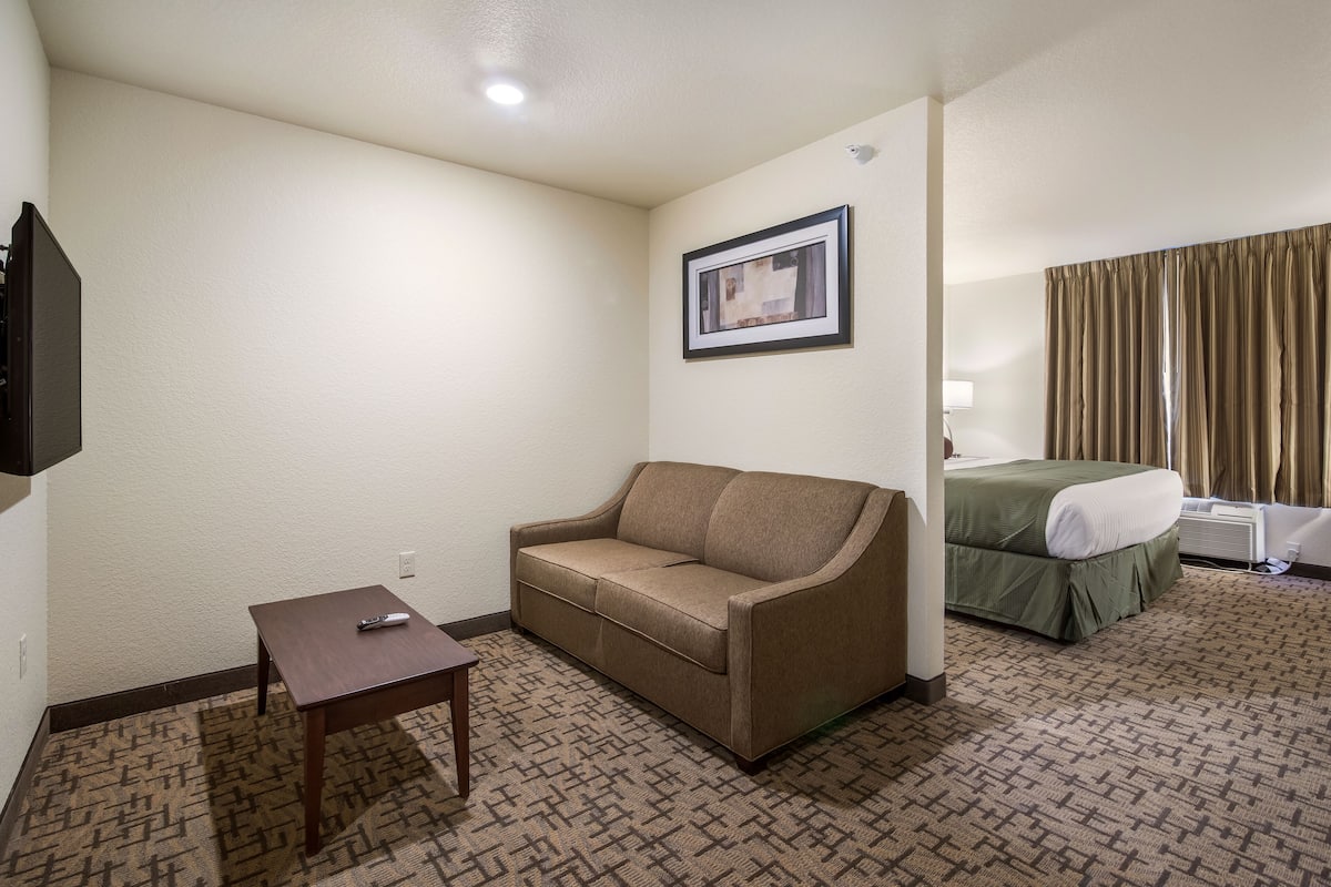 A hotel room with a sofa, a small table, a framed picture on the wall, a bed with a green bedspread, and a window with curtains.