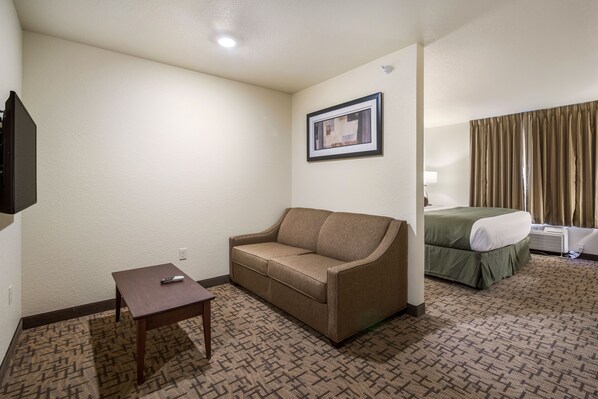 A hotel room with a sofa, a small table, a framed picture on the wall, a bed with a green bedspread, and a window with curtains.