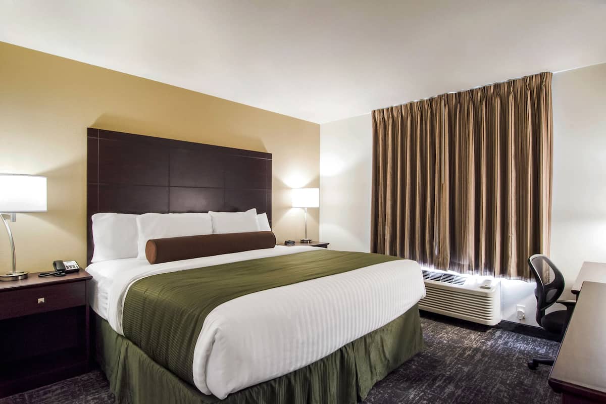 A hotel room with a large bed, two bedside lamps, a window with curtains, an air conditioning unit, and a desk with a chair.
