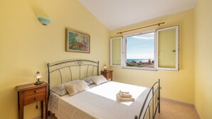A bedroom with a bed, a nightstand, a lamp, a painting, and a window with a view. - Villino Panna (Dorgali)