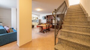 A spacious living area with a dining table and chairs, a blue sofa, and a staircase with a metal railing. - Villino Panna (Dorgali)
