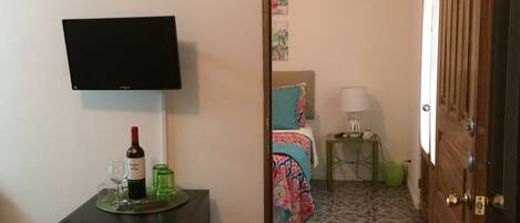 A hotel room with a bed, a bedside table with a lamp, a TV mounted on the wall, and a minibar with a bottle of wine and glasses.