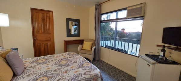 A hotel room with a bed, a chair, a small table with a bottle and glasses, a TV, and a window with a balcony view.