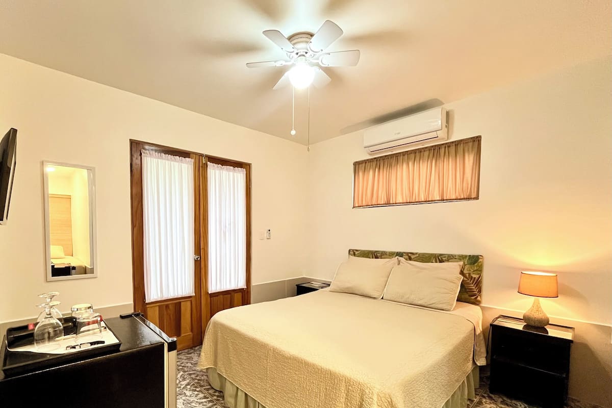 Habitación de hotel con cama, escritorio, espejo, ventilador de techo, aire acondicionado y ventana con cortinas.