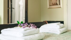 Un montón de toallas blancas dobladas con una ramita de flores moradas sobre una cama en una habitación con espejo y un cuadro en la pared.