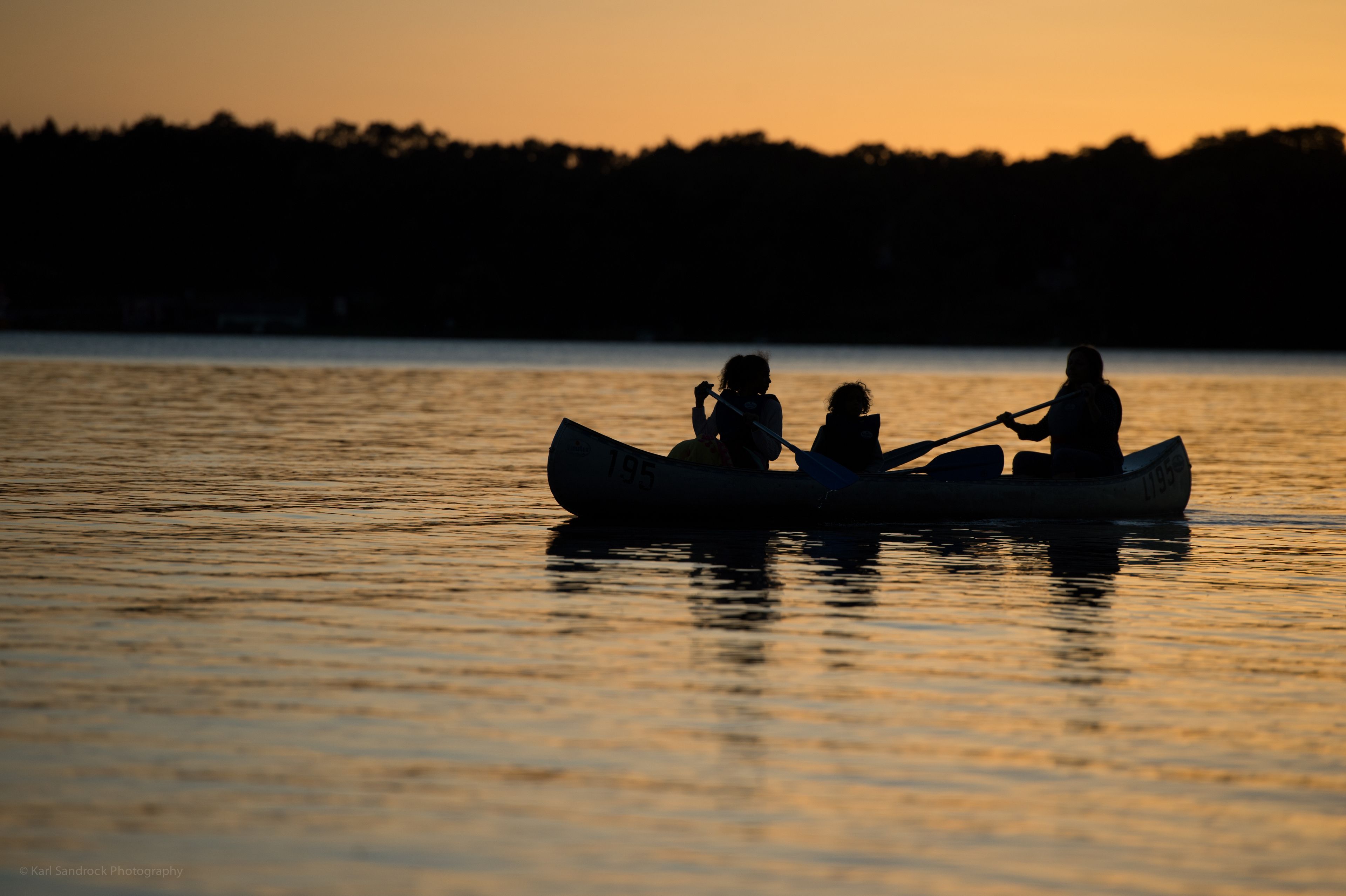 A canoe with the number 195 on it, two people paddling, and a calm water body at sunset.