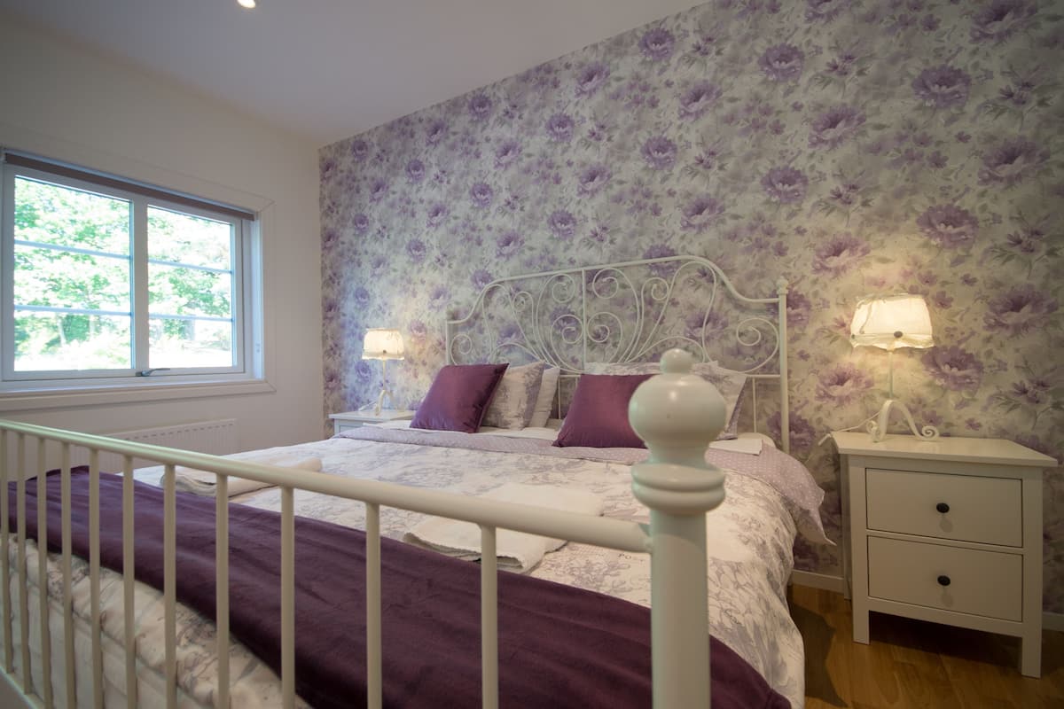 Une chambre avec un papier peint à motifs floraux, un cadre de lit en métal blanc, deux tables de chevet avec des lampes et une fenêtre avec des stores.