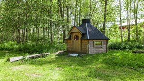 Una cabaña de madera pequeña con techo negro, rodeada de un bosque verde denso.