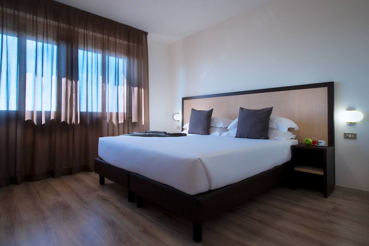 A neatly made bed with a wooden headboard, a bedside table with a lamp, and a window with curtains.