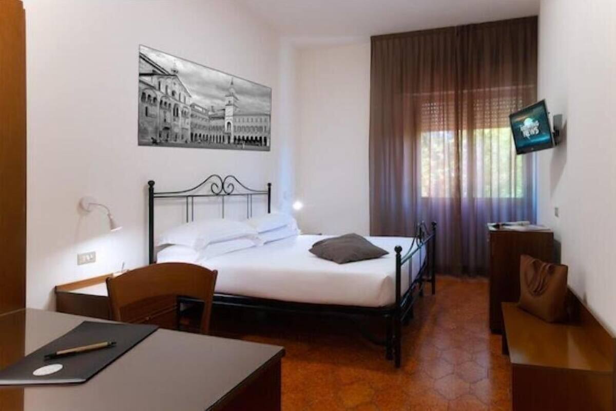 A hotel room with a bed, a desk, a chair, a TV, and a painting on the wall.