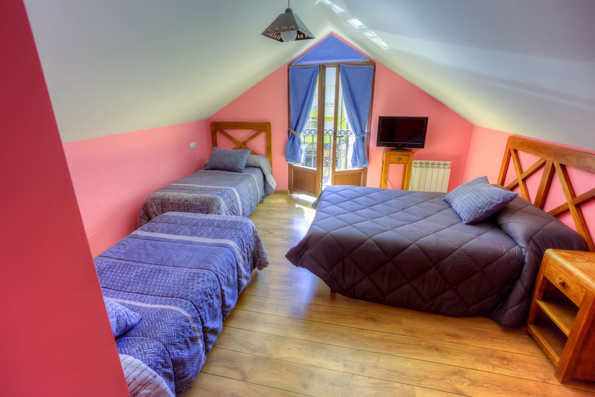 A cozy attic bedroom with two beds, a television, and a window with blue curtains.